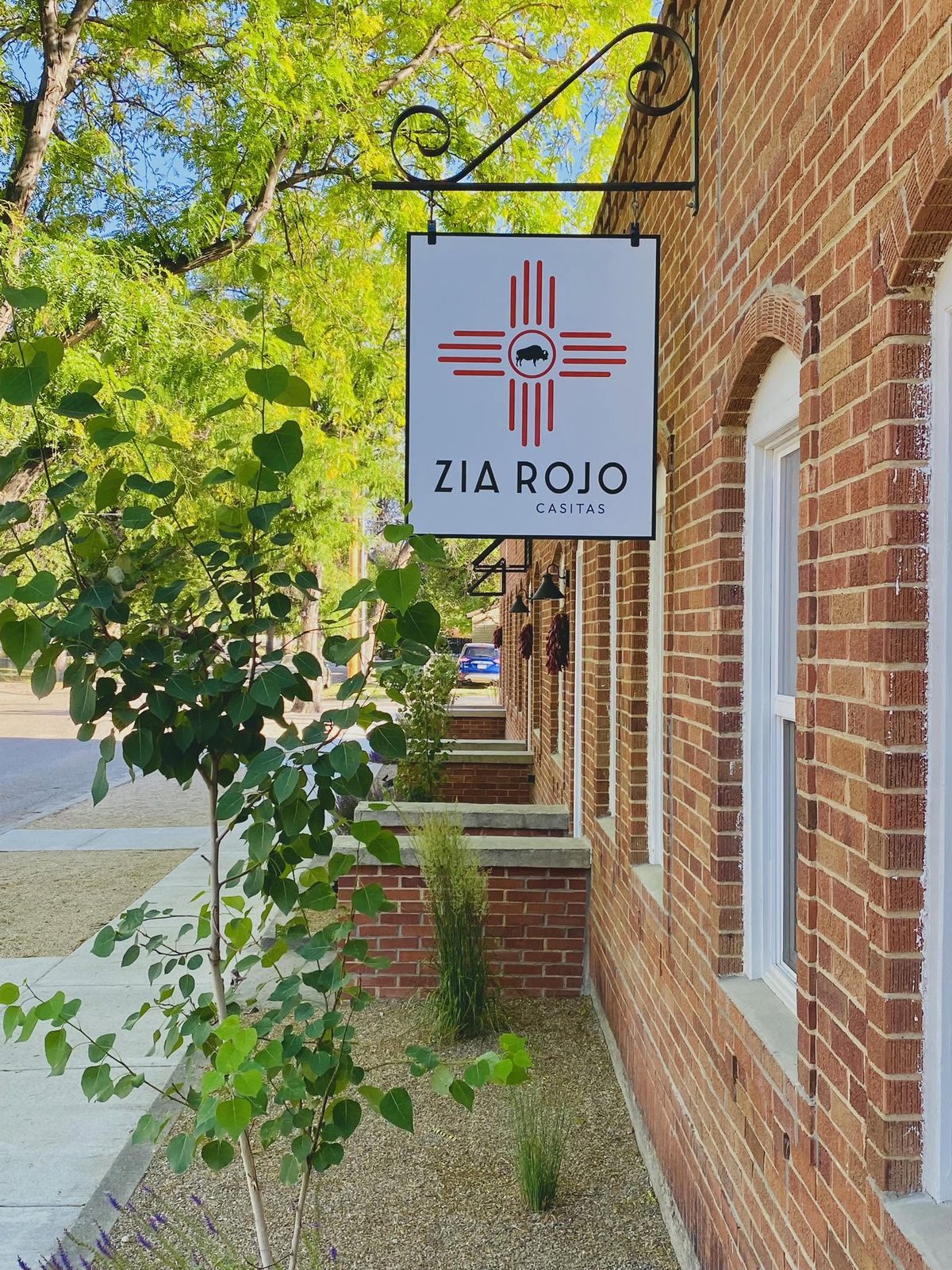Zia Rojo Casitas hanging sign on a red-brick building beside a tree-lined sidewalk