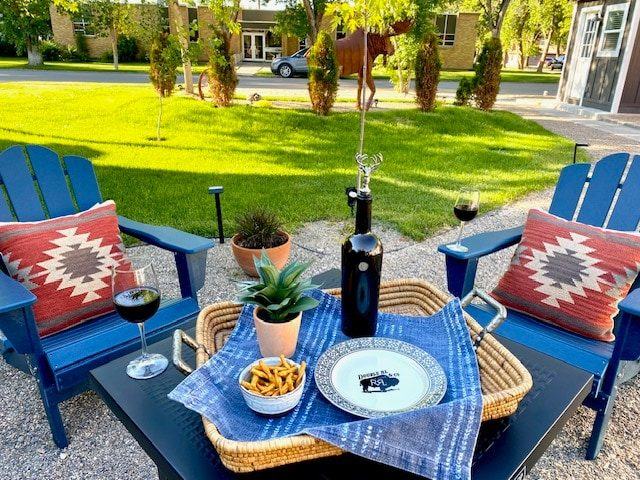 Blue Adirondack chairs and a table with red patterned pillows, a basket and wine on the lawn